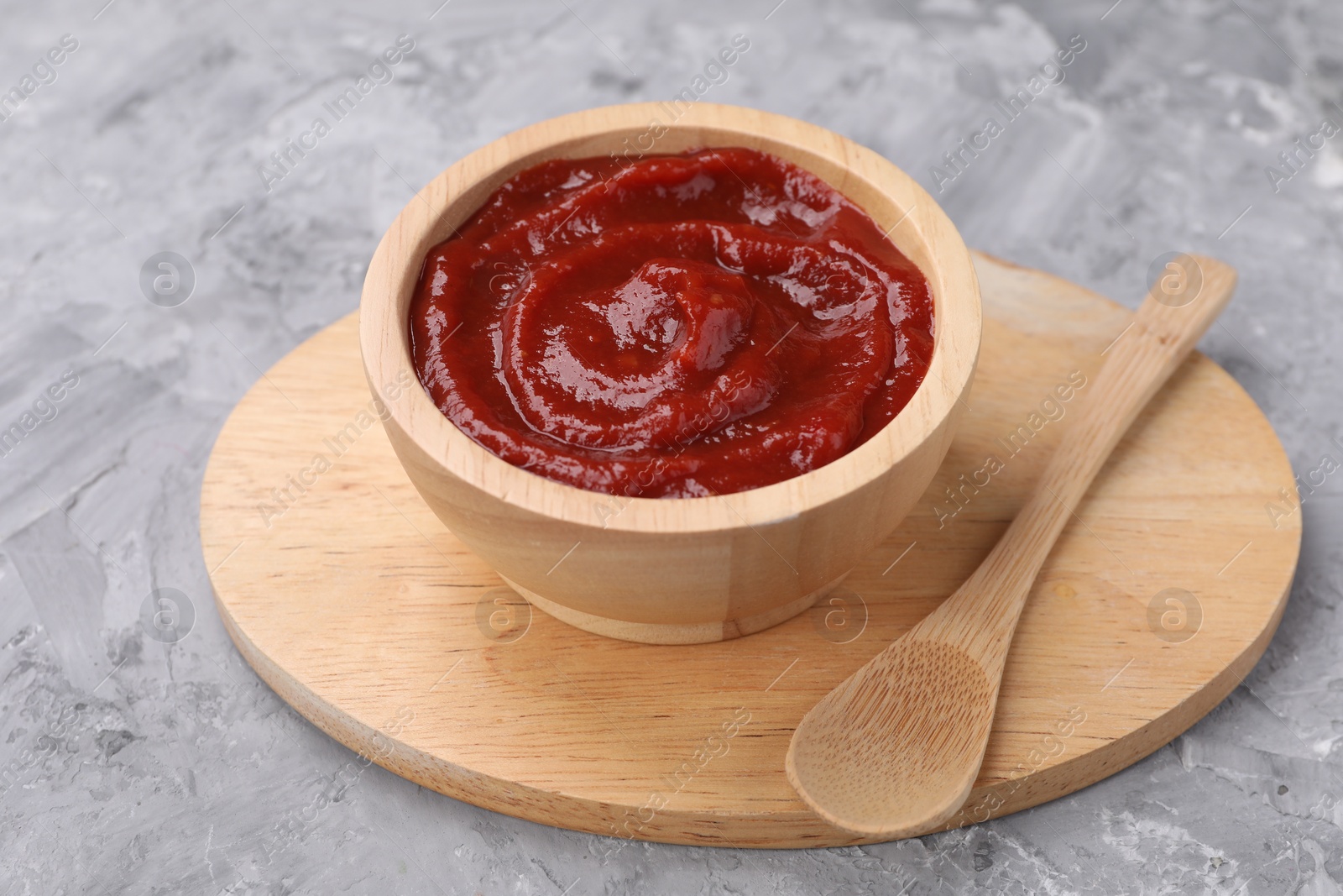 Photo of Organic ketchup in wooden bowl and spoon on grey textured table. Tomato sauce