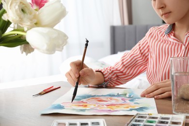 Woman painting flowers with watercolor at wooden table indoors, closeup. Creative artwork