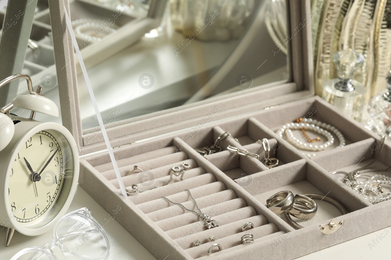 Photo of Jewelry box with many different accessories and alarm clock on white table, closeup