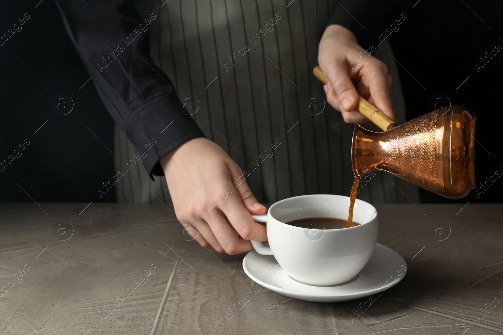 Photo of Turkish coffee. Woman pouring brewed beverage from cezve into cup at gray table against black background, closeup