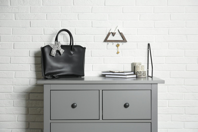 Grey chest of drawers with bag, books and decor elements near white brick wall