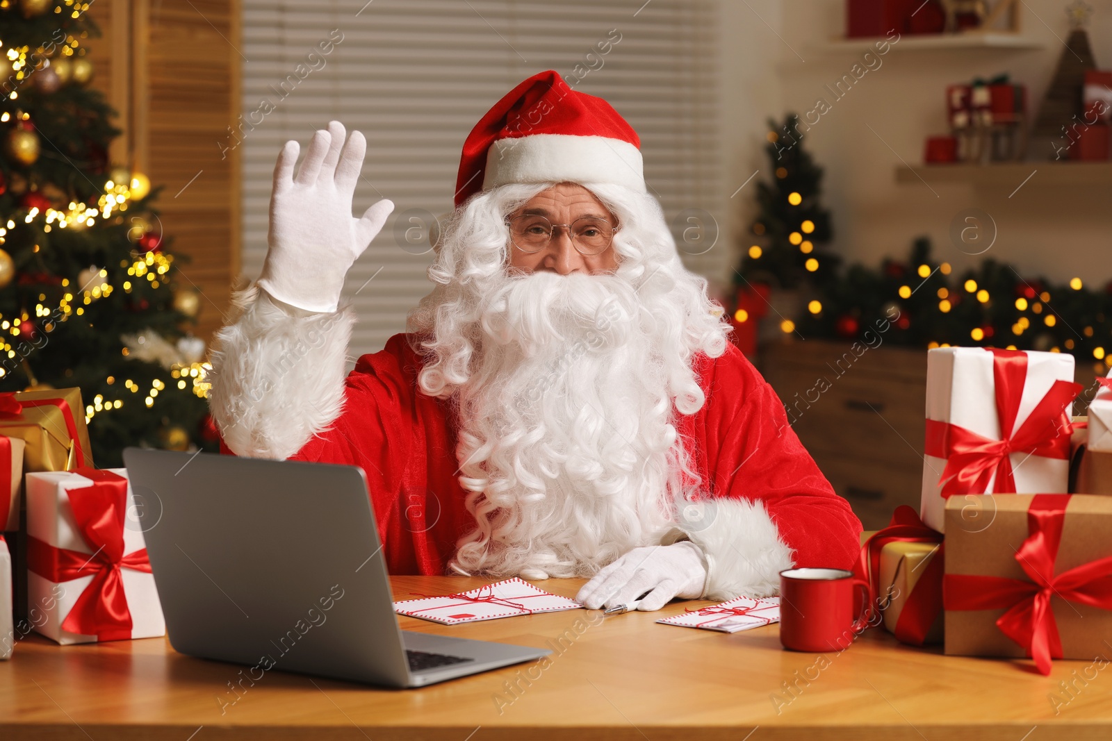Photo of Santa Claus raising hand. Laptop, Christmas gifts and letters on table