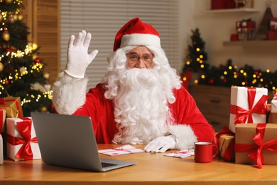 Santa Claus raising hand. Laptop, Christmas gifts and letters on table