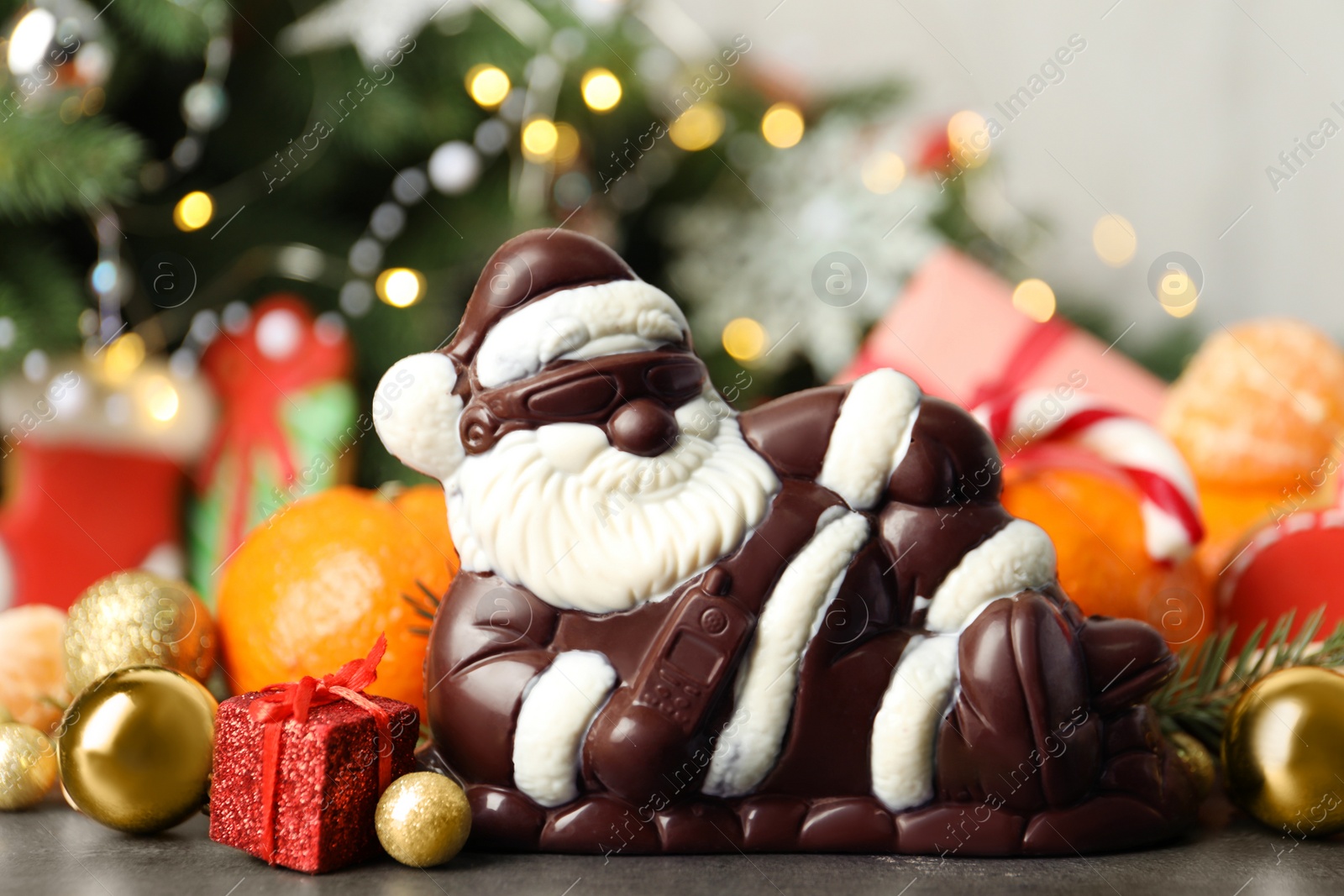 Photo of Composition with chocolate Santa Claus among sweets, decorations and tangerine fruits against Christmas tree, closeup