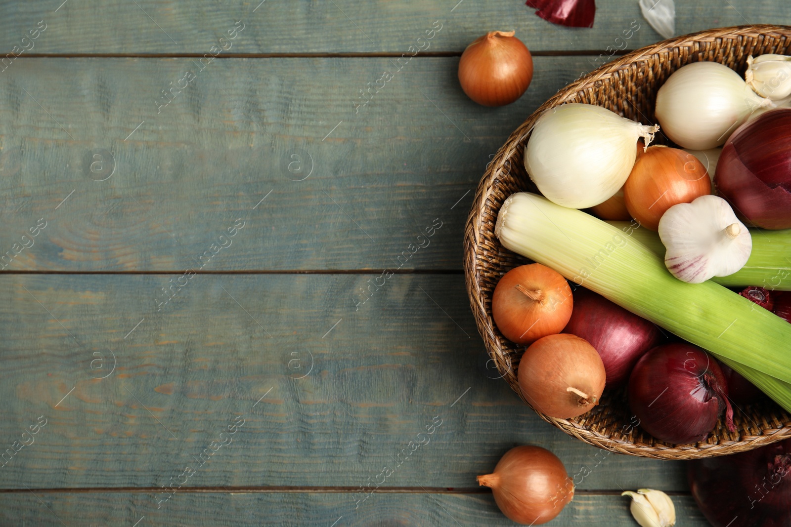 Photo of Wicker basket with fresh onion bulbs, leeks and garlic on light blue wooden table, flat lay. Space for text