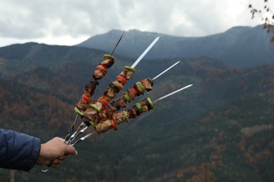 Photo of Man holding metal skewers with delicious meat and vegetables against mountain landscape, closeup