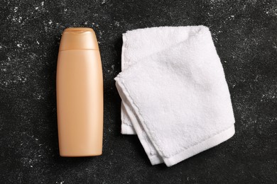 Photo of Soft folded terry towel and cosmetic bottle on black textured background, flat lay
