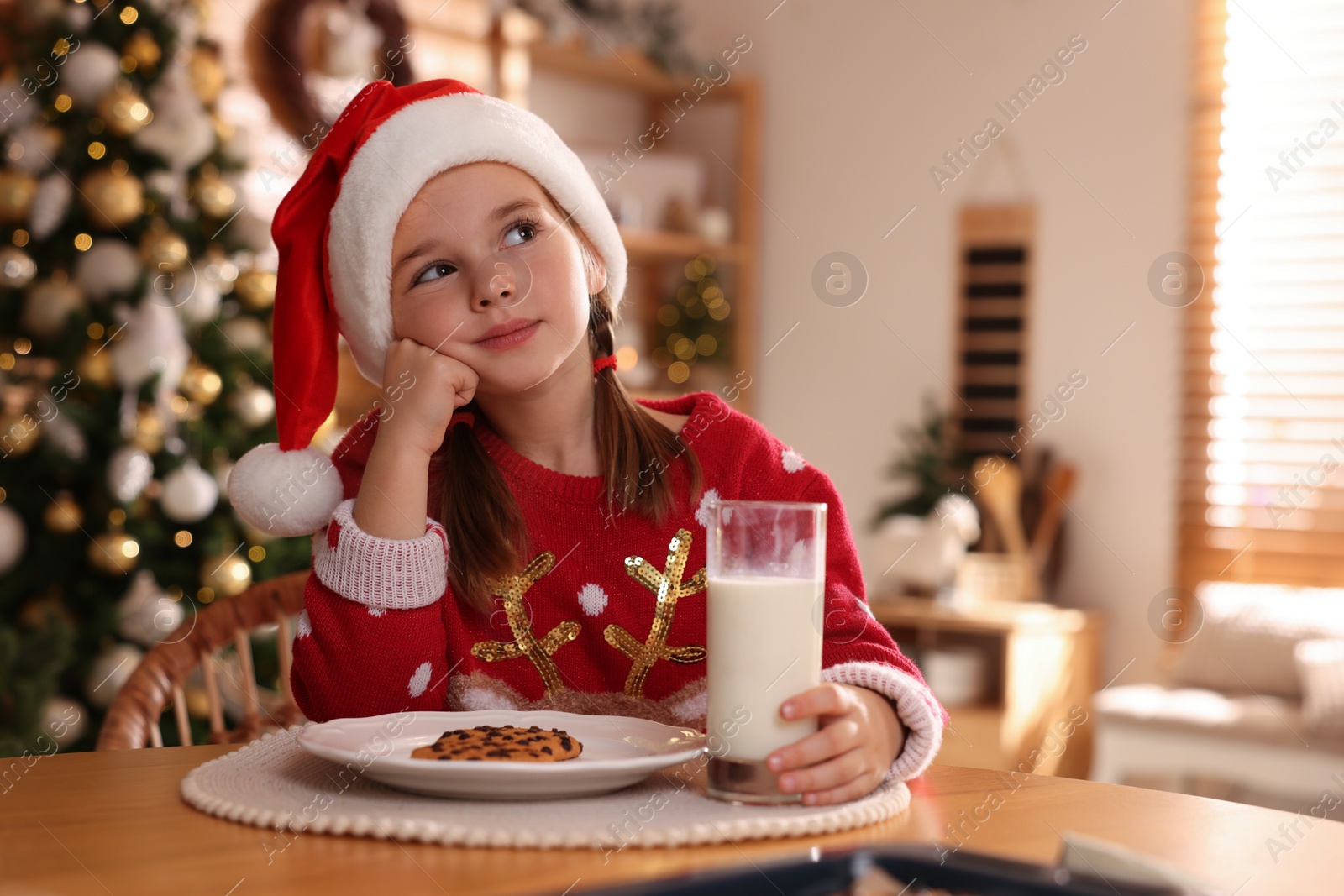 Photo of Cute little girl in Santa hat with delicious Christmas cookie and milk at home, space for text