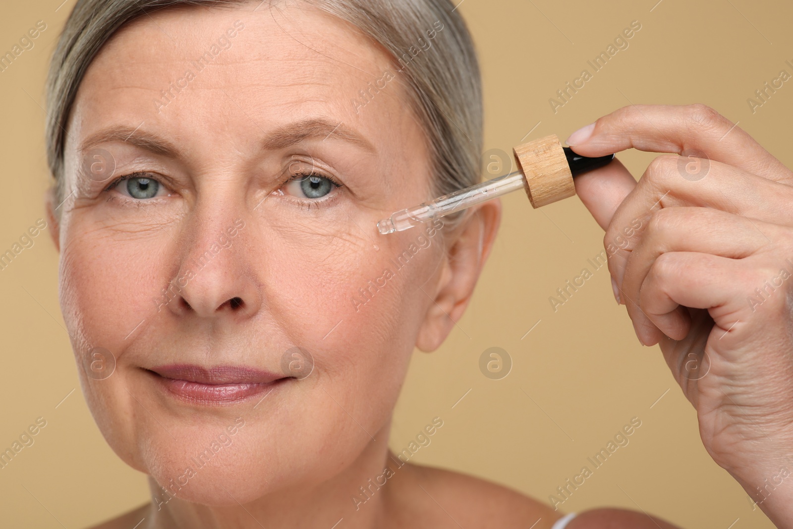 Photo of Senior woman applying cosmetic product on her aging skin against beige background. Rejuvenation treatment