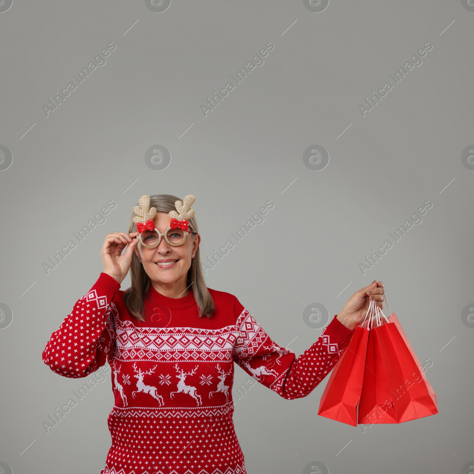 Photo of Happy senior woman in Christmas sweater and funny glasses with shopping bags on grey background