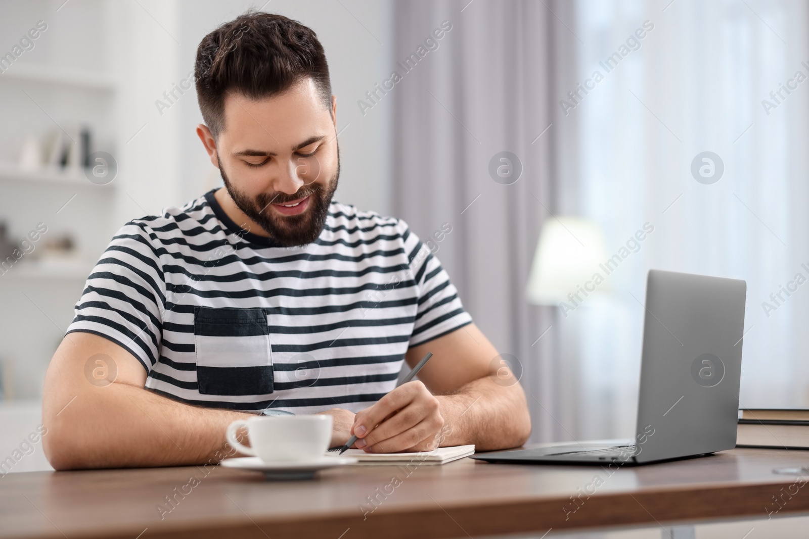 Photo of Young man writing down notes during webinar at table in room