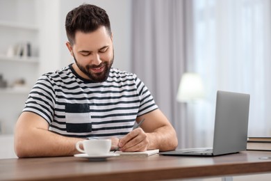 Photo of Young man writing down notes during webinar at table in room