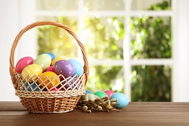 Image of Colorful Easter eggs in wicker basket and willow branches on wooden table, space for text
