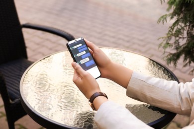 Woman texting via mobile phone at table outdoors, closeup. Device screen with messages