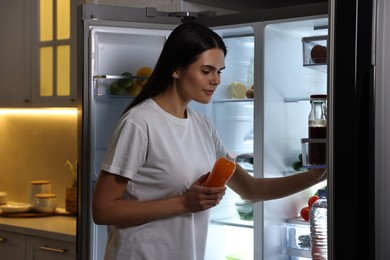 Photo of Young woman taking bottle of juice out of refrigerator in kitchen at night