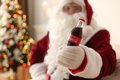 Photo of MYKOLAIV, UKRAINE - JANUARY 18, 2021: Santa Claus with Coca-Cola bottle in room decorated for Christmas, focus on hand