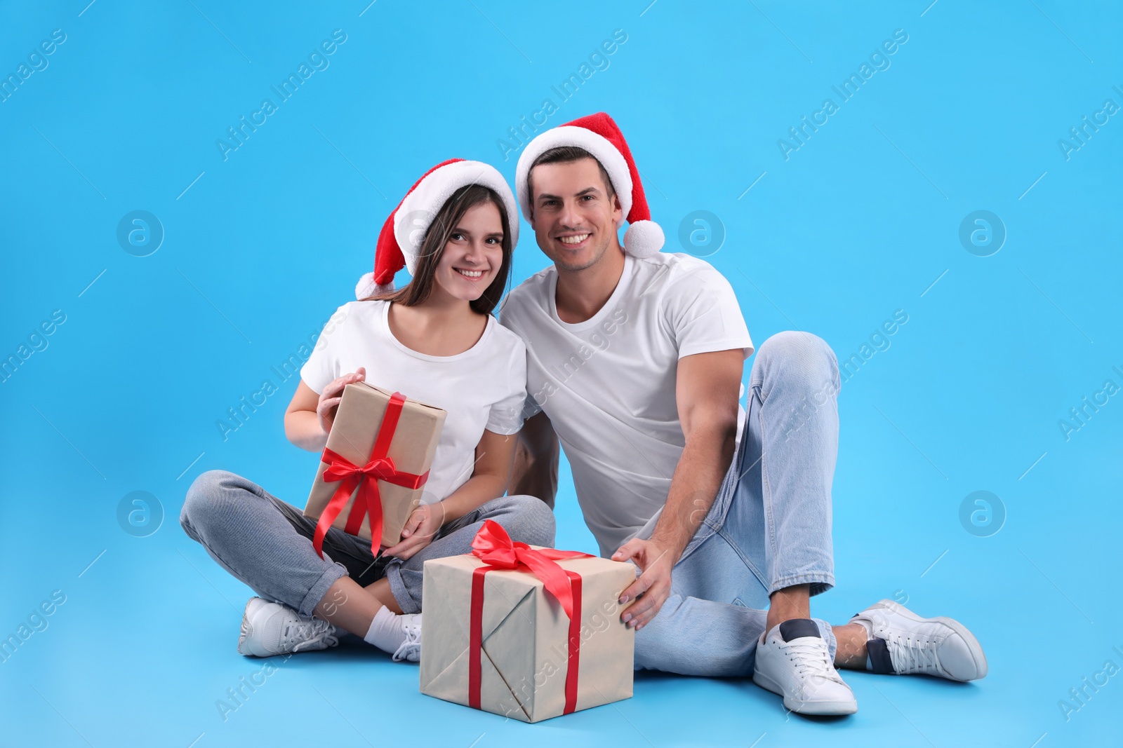 Photo of Beautiful happy couple in Santa hats sitting with Christmas gifts on light blue background