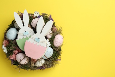 Photo of Wooden bunnies with protective masks, painted eggs, Easter wreath and space for text on yellow background, top view. Holiday during COVID-19 quarantine