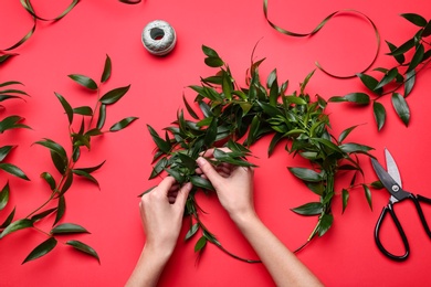 Photo of Florist making beautiful mistletoe wreath on red background, top view. Traditional Christmas decor