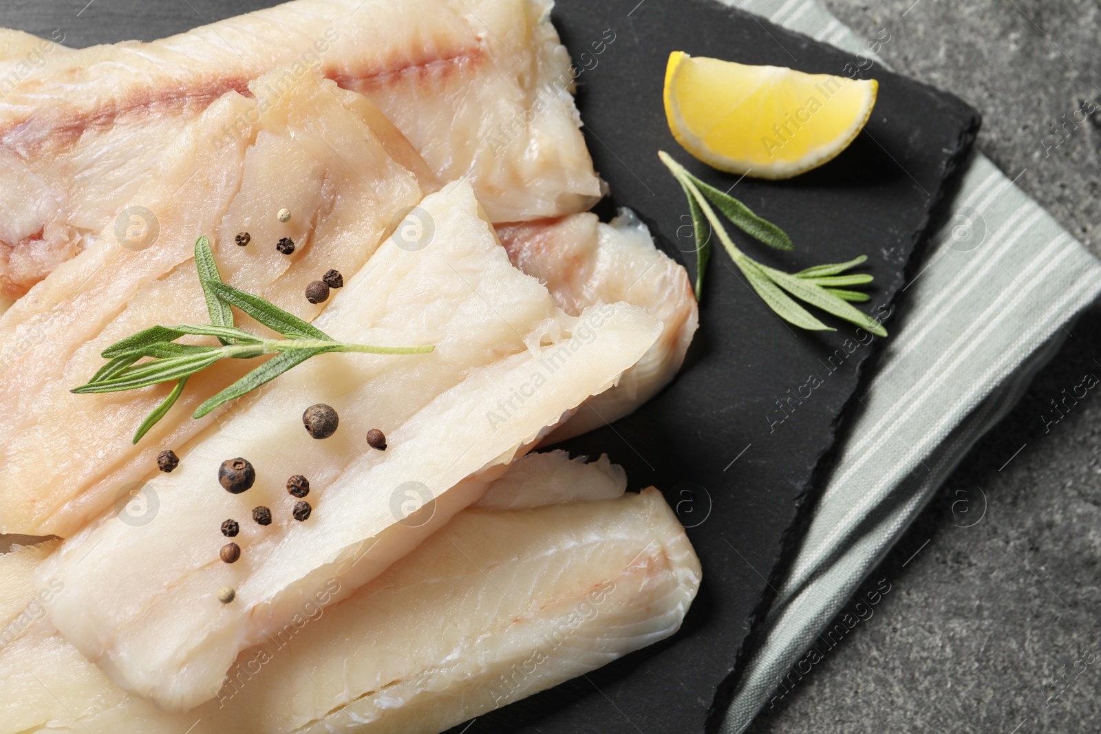 Photo of Pieces of raw cod fish, rosemary, peppercorns and lemon on grey table, closeup