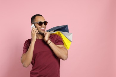 Photo of Happy African American man in sunglasses with shopping bags talking on smartphone against pink background. Space for text