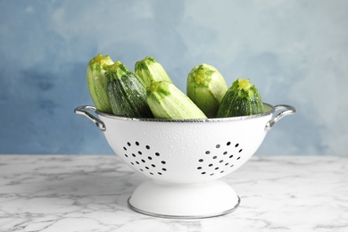 Photo of Colander with fresh ripe green zucchinis on marble table against blue background