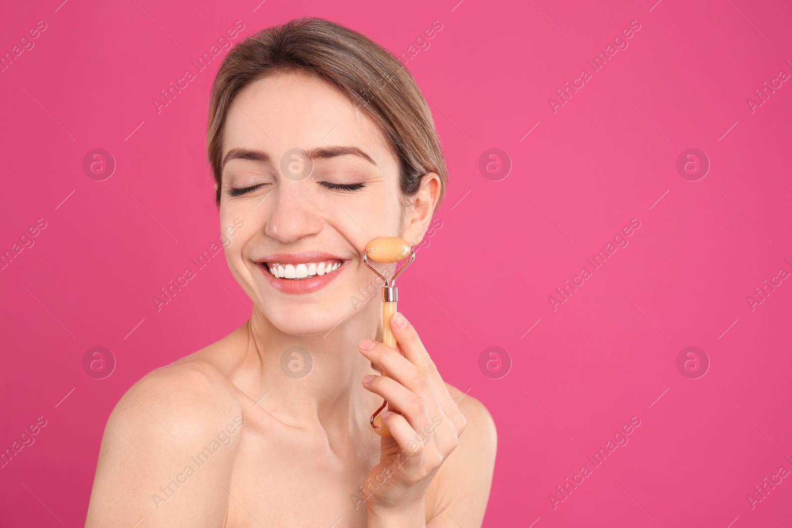 Photo of Young woman using natural jade face roller on pink background, space for text