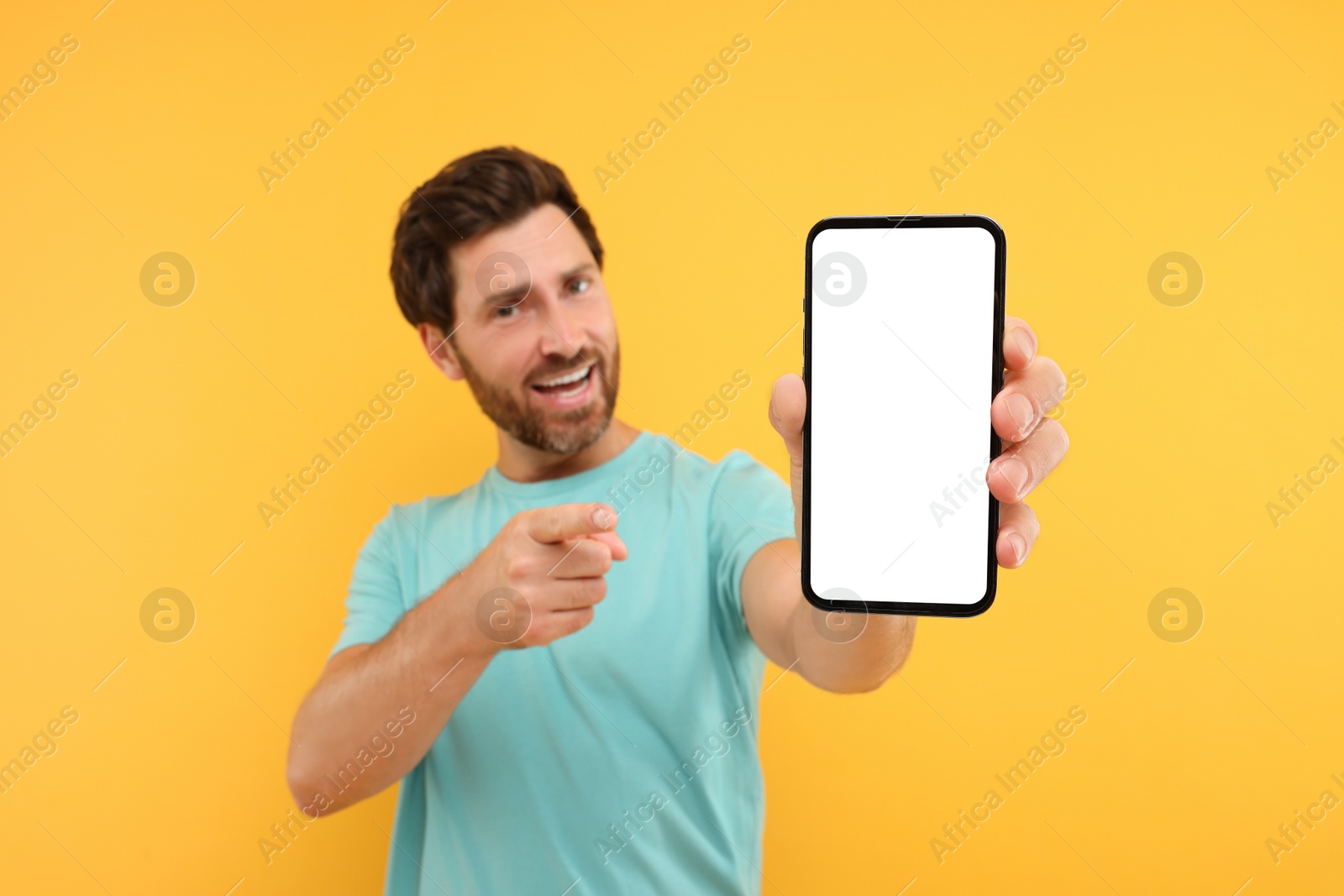 Photo of Handsome man showing smartphone in hand and pointing at it on yellow background, selective focus. Mockup for design