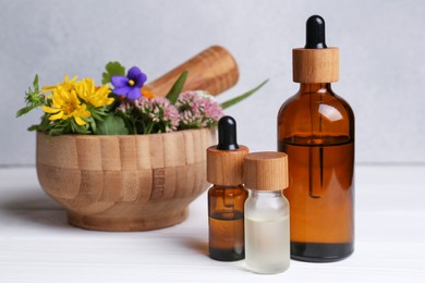Photo of Glass bottles of aromatic essential oil, mortar with different wildflowers on white wooden table, closeup