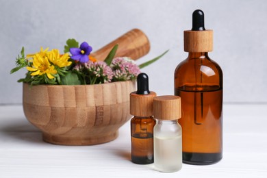 Glass bottles of aromatic essential oil, mortar with different wildflowers on white wooden table, closeup