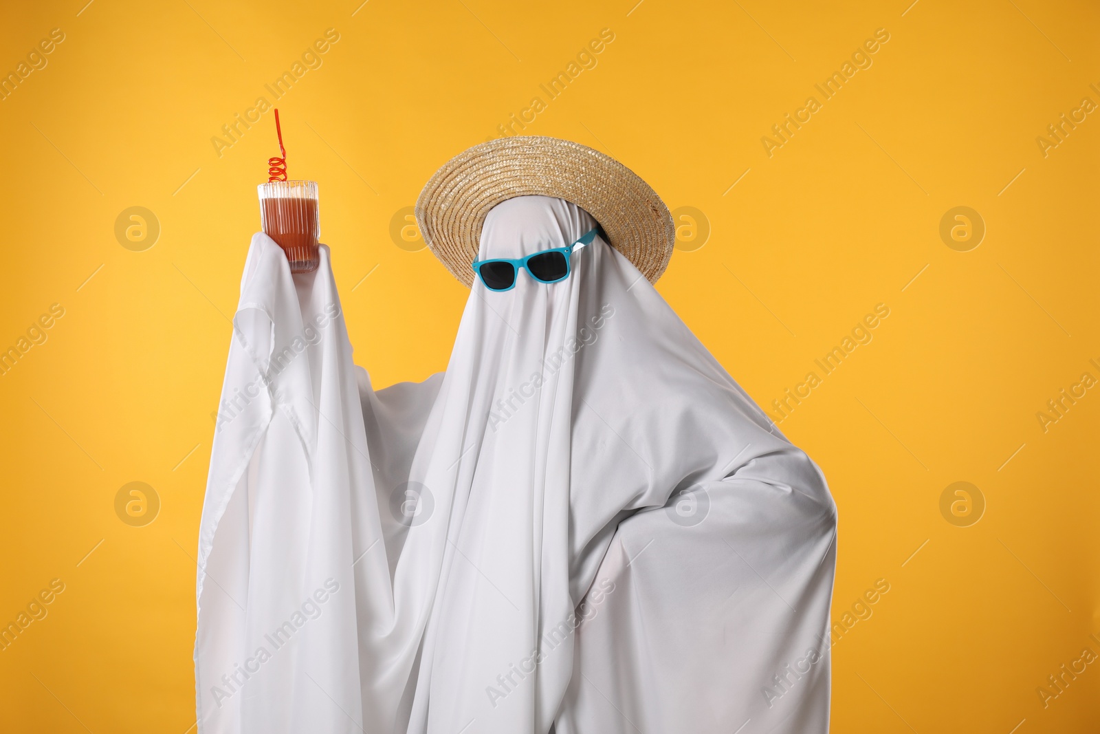 Photo of Person in ghost costume, sunglasses and straw hat holding glass of drink on yellow background