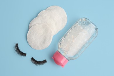 Bottle of makeup remover, cotton pads and false eyelashes on light blue background, flat lay