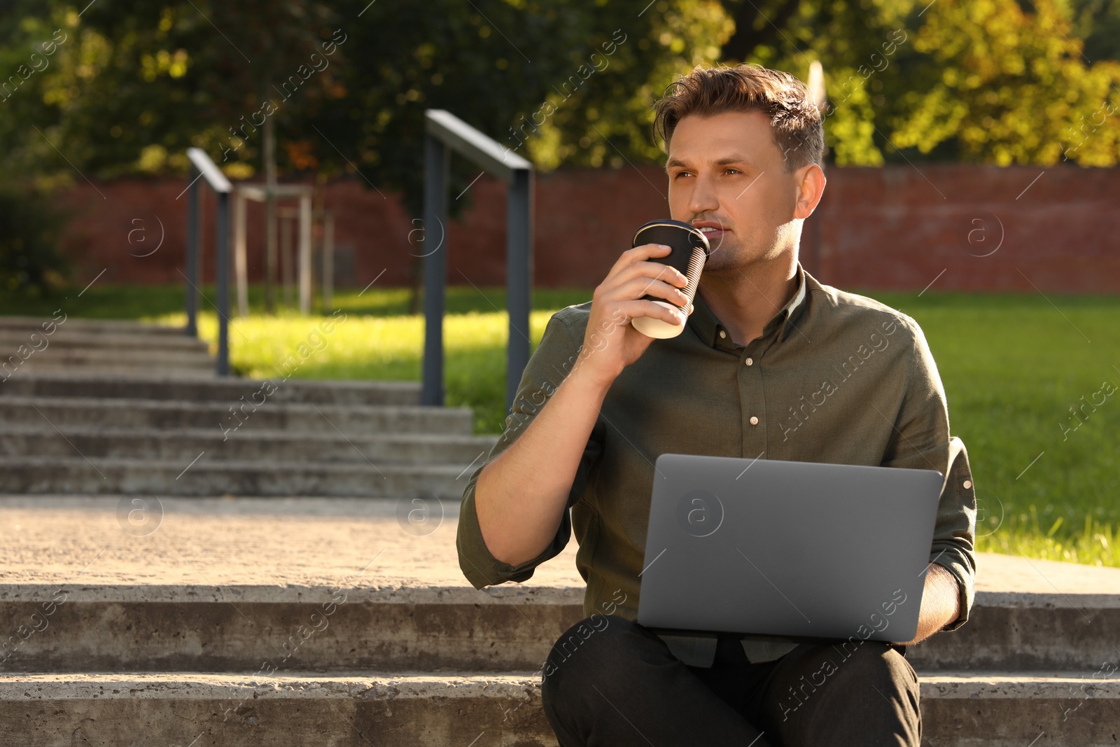 Photo of Handsome man with laptop drinking coffee on stairs outdoors. Space for text