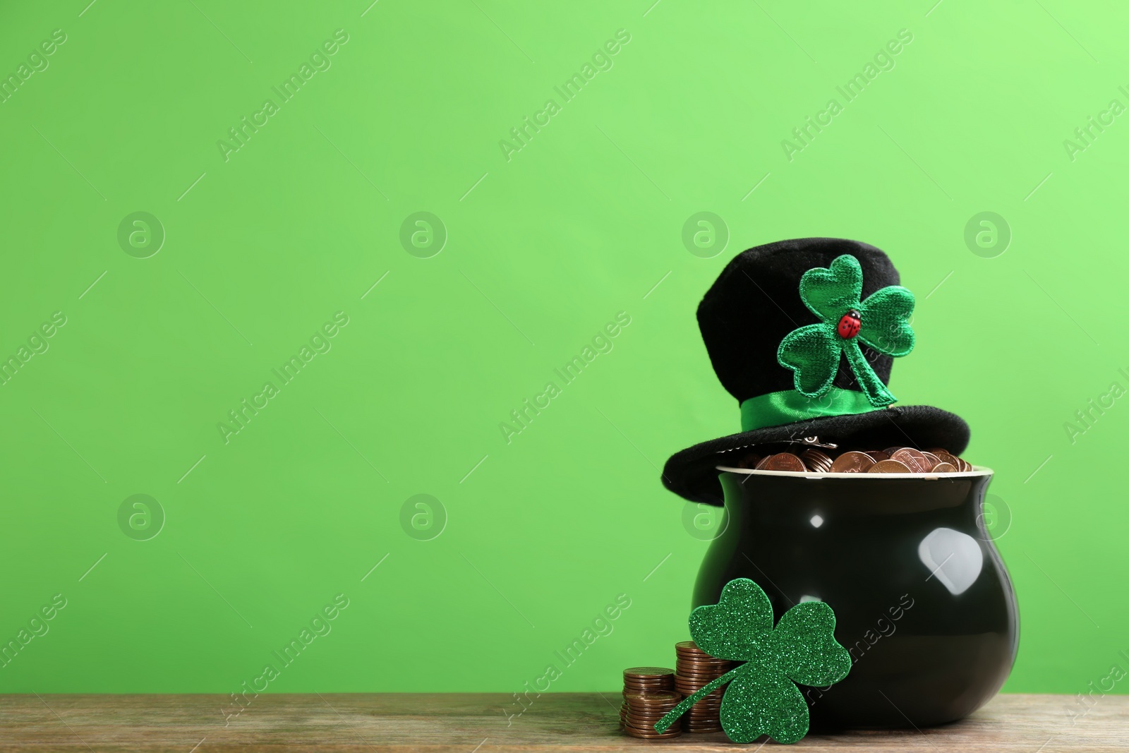 Photo of Pot of gold coins, hat and clover on wooden table against green background, space for text. St. Patrick's Day celebration