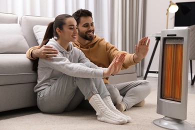 Photo of Young couple warming hands near electric heater at home