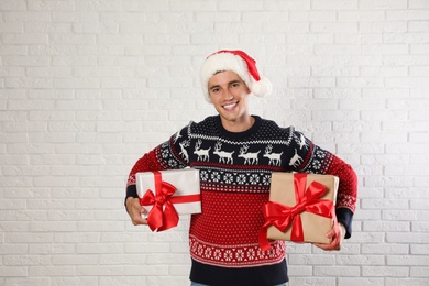 Photo of Happy man in Christmas sweater and Santa hat holding gift boxes near white brick wall