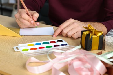 Man writing message in greeting card at wooden table, closeup