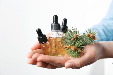 Woman holding bottles of essential oils and fir branches on white background, closeup