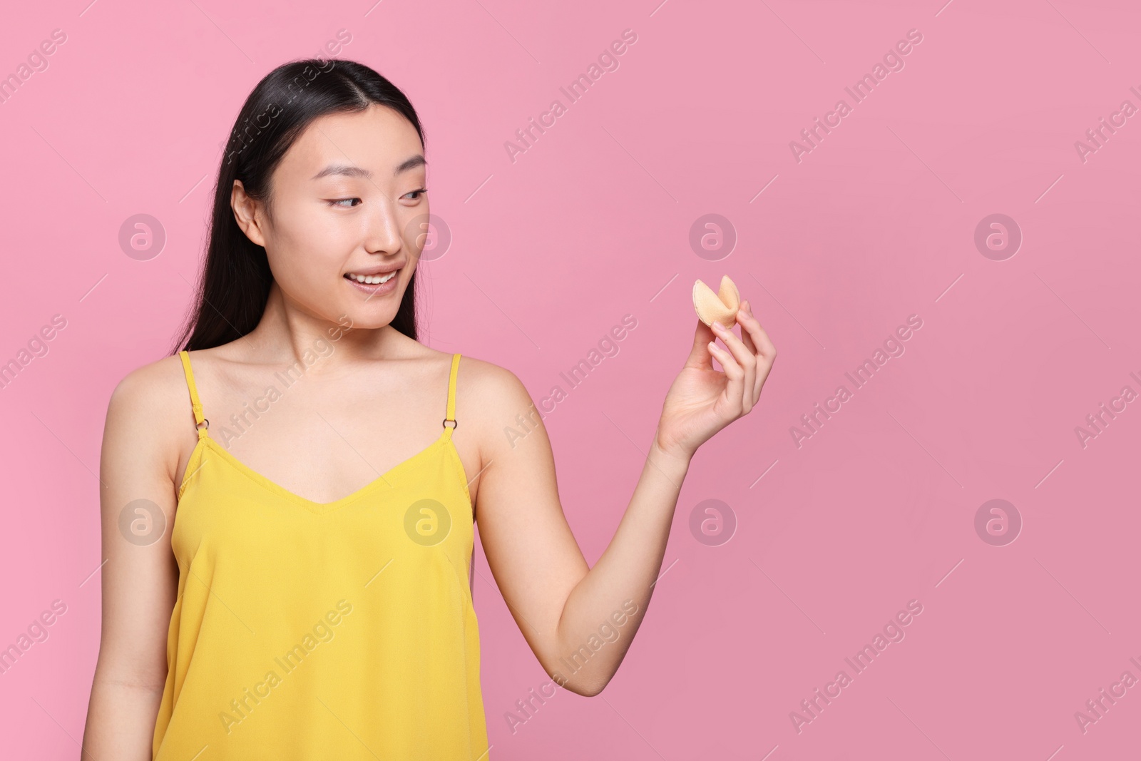 Photo of Asian woman holding tasty fortune cookie with prediction on pink background, space for text