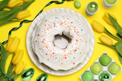 Photo of Easter cake with sprinkles, painted eggs and tulips on yellow background, flat lay