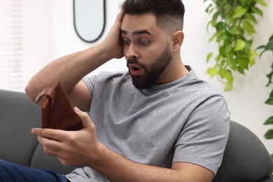 Confused man with empty wallet on sofa indoors