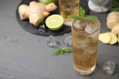 Photo of Glass of tasty ginger ale with ice cubes and ingredients on grey wooden table, closeup. Space for text