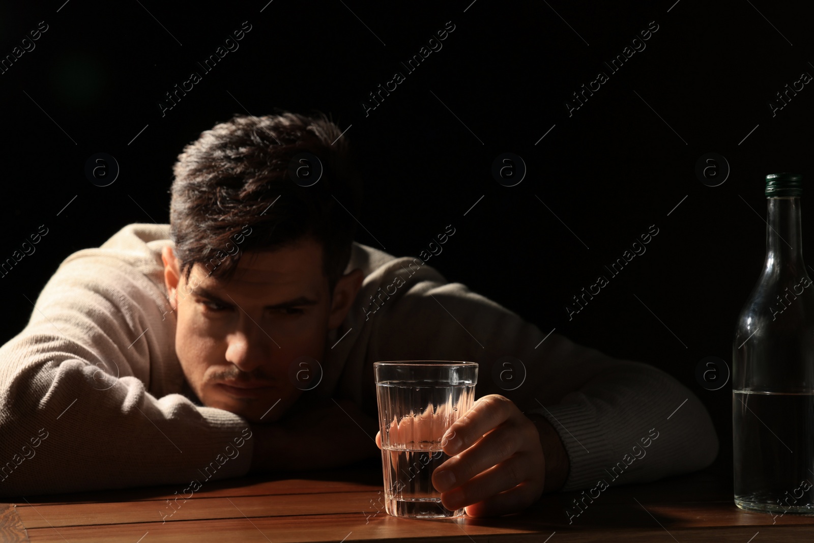 Photo of Addicted man with alcoholic drink at wooden table against black background, focus on glass