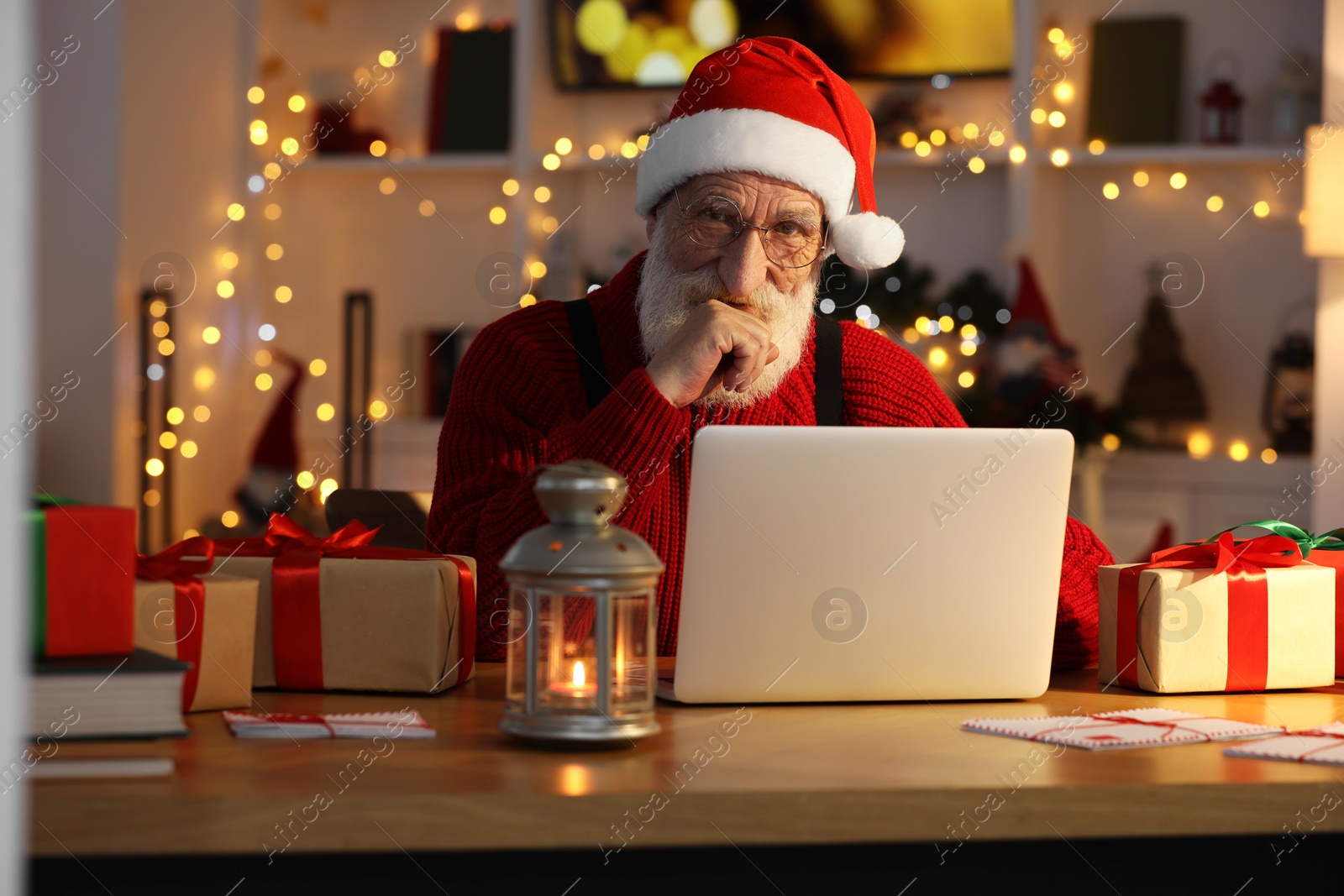 Photo of Santa Claus using laptop at his workplace in room decorated for Christmas