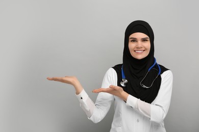 Photo of Muslim woman in hijab and medical uniform pointing at something on light gray background, space for text