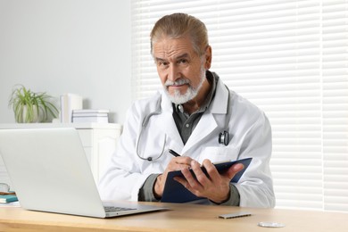 Photo of Senior doctor with laptop and clipboard consulting patient at wooden desk in clinic. Online medicine