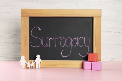 Photo of Small chalkboard with word Surrogacy, family figure and cubes on pink wooden table