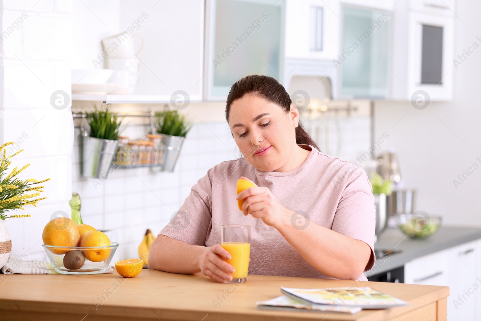 Photo of Overweight woman squeezing orange juice into glass at table in kitchen. Healthy diet