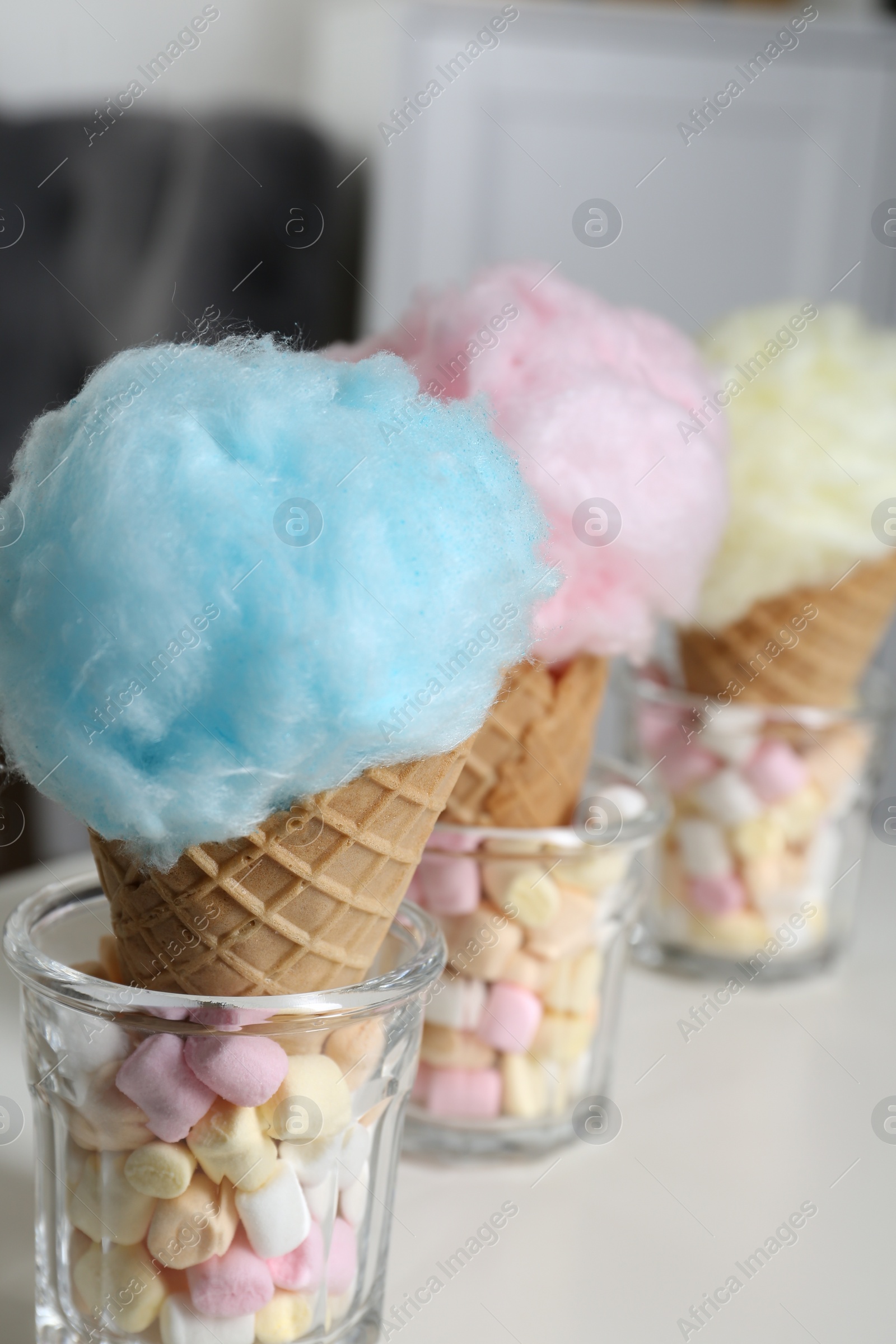 Photo of Sweet cotton candies in waffle cones and marshmallows on white table indoors, closeup