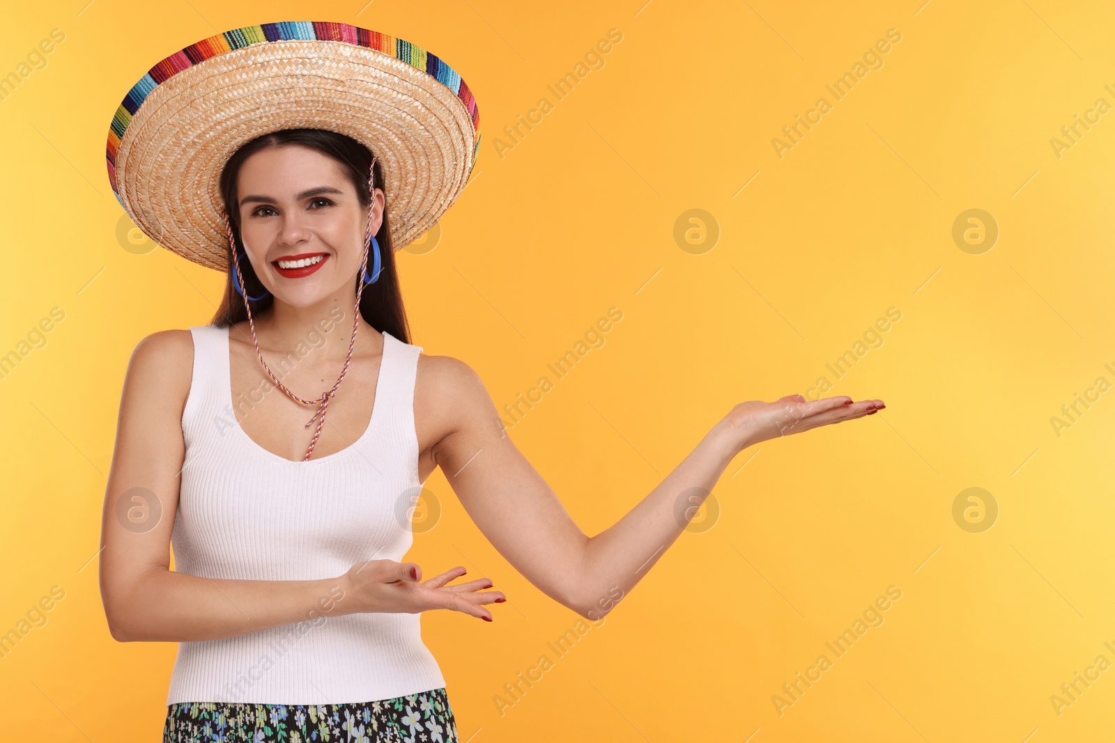 Photo of Young woman in Mexican sombrero hat showing something on yellow background. Space for text